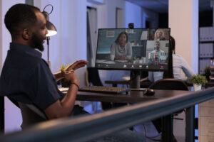 Executive manager in a modern office setting engaged in a virtual meeting with a diverse team. The image highlights the use of video conferencing technology for effective communication and collaboration, with participants displayed on a computer screen. The office features contemporary decor and dim lighting, emphasizing a professional and connected work environment.