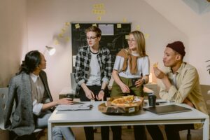 A group of four coworkers gathered around a desk in an office, chatting and eating pizza during a casual break, with a chalkboard and sticky notes in the background.